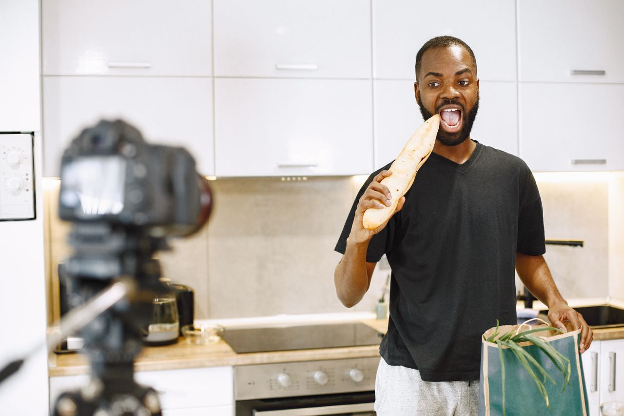 who-we-are Un homme filme son quotidien dans sa cuisine avec une caméra, tenant une baguette et un sac de courses.